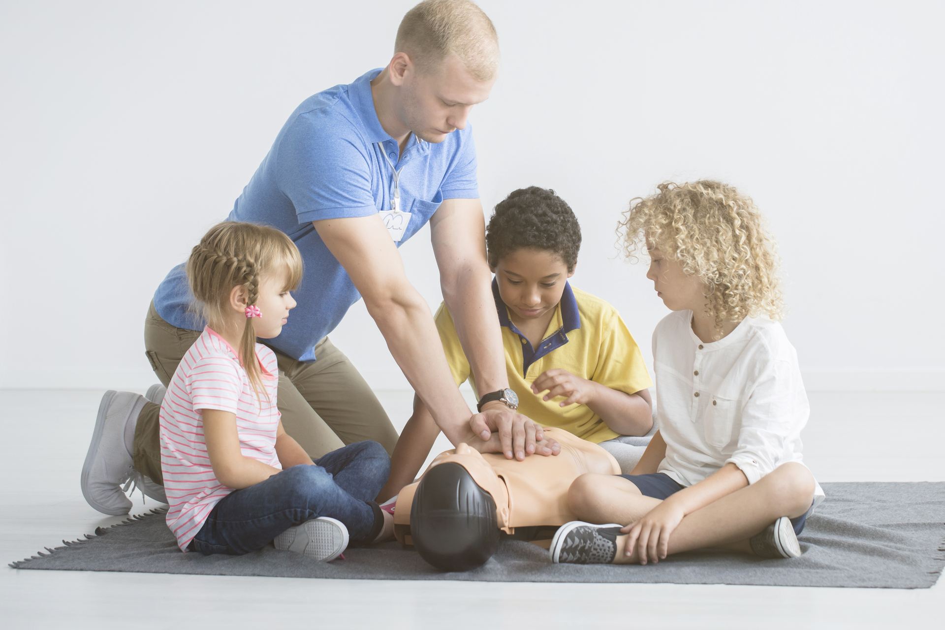 Ein Ausbilder zeigt drei Grundschulkindern die Herzdruckmassage an einer Übungspuppe